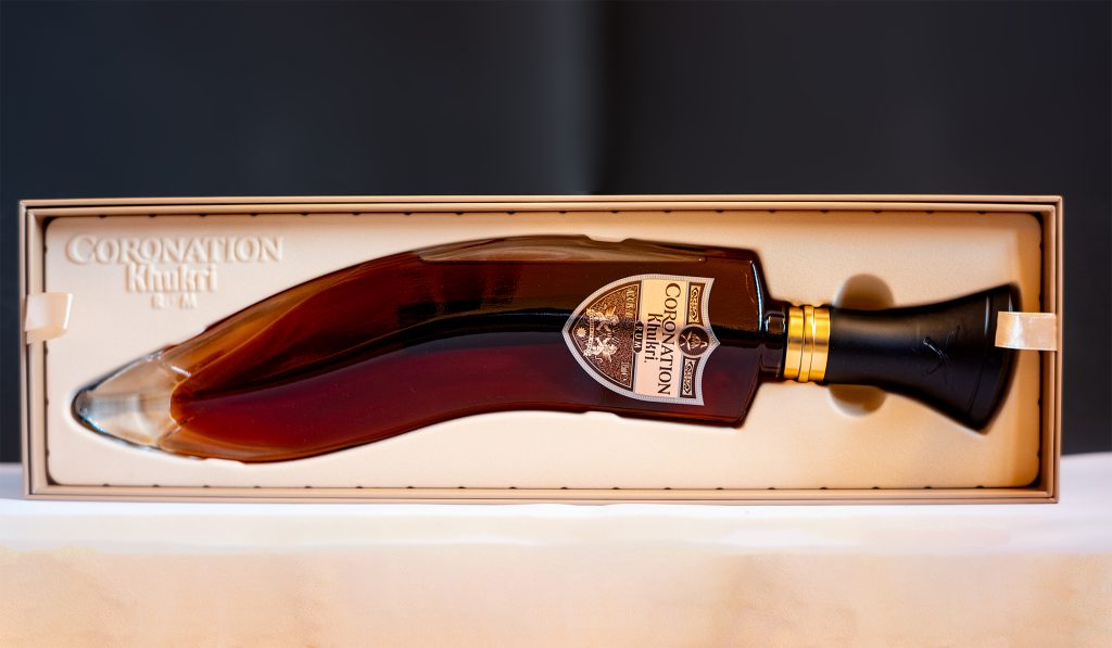 Two-part image showing Khukri Coronation Rum in a velvet-lined display box above its ornate black textured presentation case.

